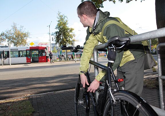 Ein junger mann schließt sein Fahrrad an einem Fahrradbügel an und schaut zu einer Straßenbahn im Hintergrund. 